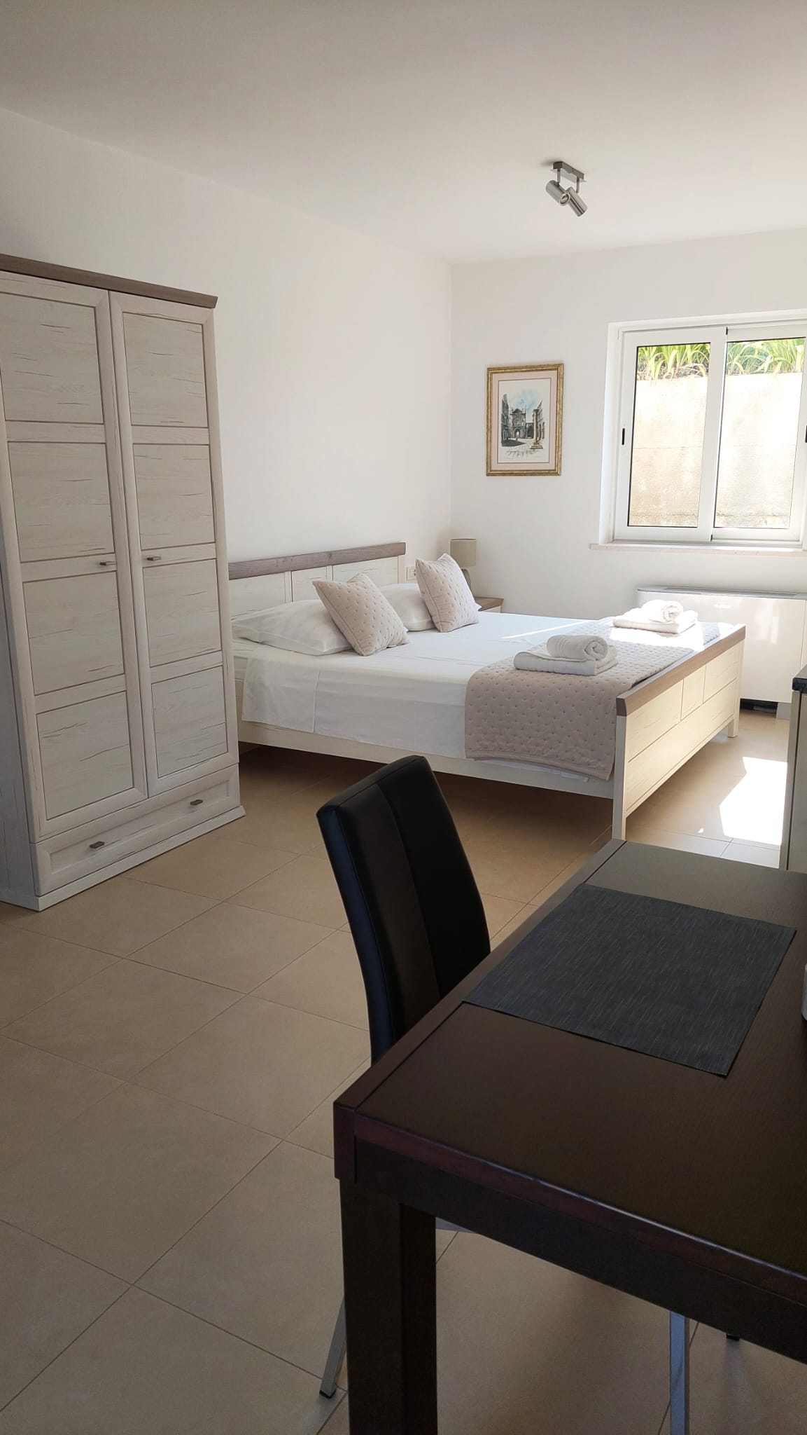 A well-lit bedroom features a spacious bed with crisp linens and decorative pillows. A large wardrobe provides storage and a workspace with a dark chair is positioned beside the bed. Natural light enters through the window, illuminating the neutral color scheme.