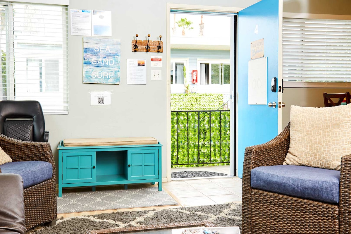 A welcoming living area features two comfortable chairs with soft cushions. A teal bench offers storage and seating. The door is open, revealing a glimpse of a green balcony. Natural light filters through the windows, highlighting the neutral-toned walls and patterned carpet.