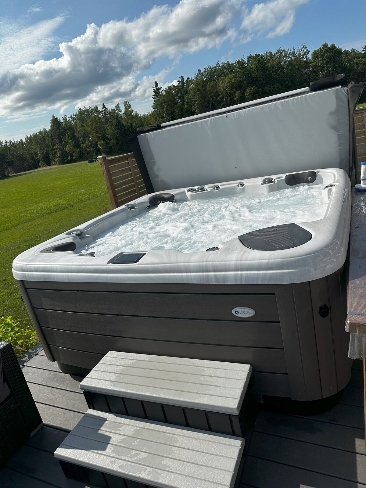 A spacious hot tub is situated on a wooden deck, with steps leading up to it. The tub is filled with bubbling water and features a gray cover partially opened. A green lawn and tree line are visible in the background under a partly cloudy sky.