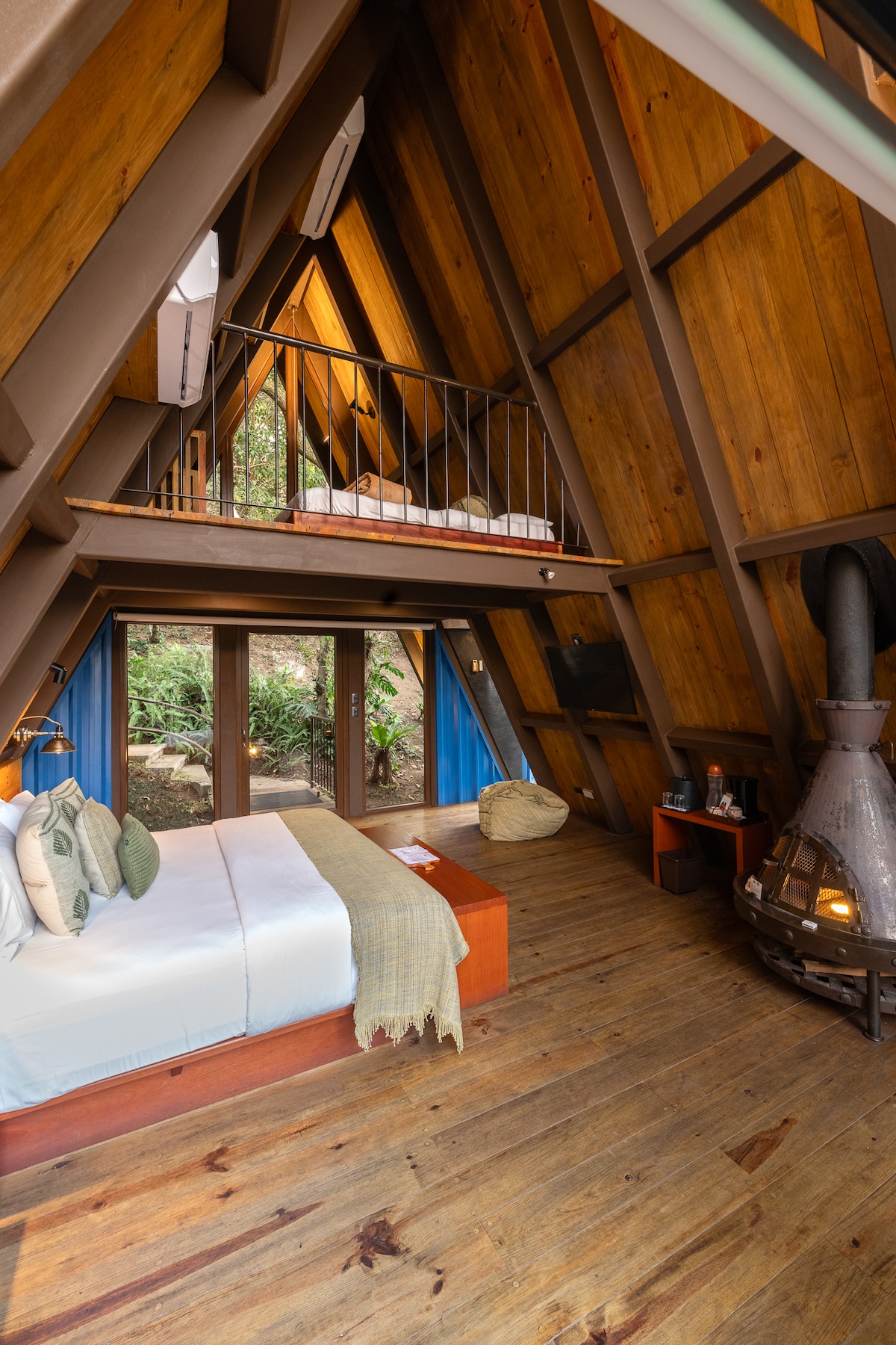 A split-level cabin features a spacious bed adorned with neutral linens. Wood-paneled walls create a warm ambiance. A cozy loft area with a railing is visible above. Large glass doors invite natural light, revealing greenery outside. A sleek fireplace adds a touch of comfort.