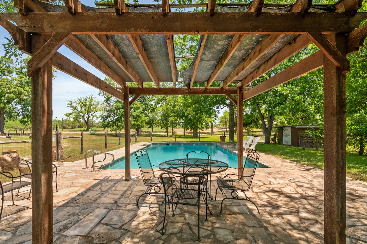 A spacious outdoor patio area features a stone floor and a sturdy wooden pergola, providing shade over a circular table and several metal chairs. The inviting swimming pool is visible in the background, surrounded by a grassy area and trees.