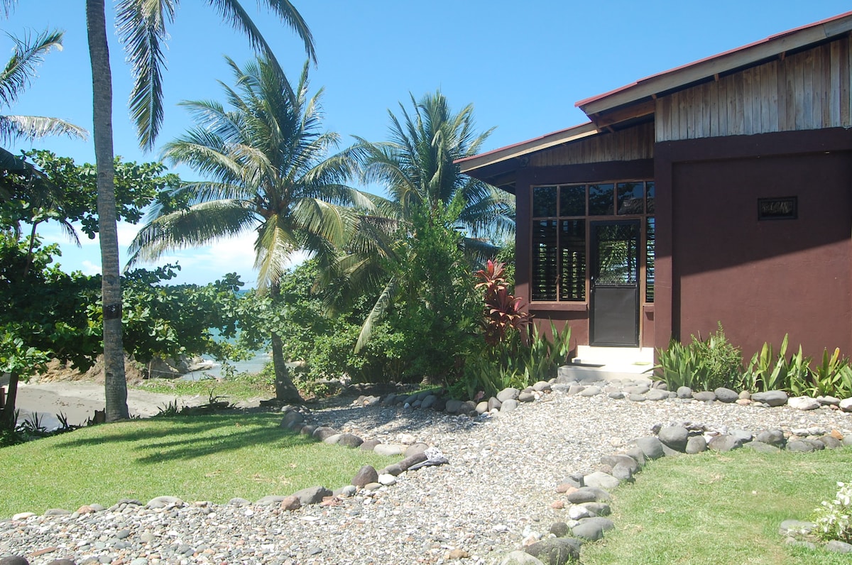 A peaceful entrance of a brown exterior unit is highlighted by paths of smooth stones and lush greenery. Palm trees gently sway nearby, with a view of the ocean visible in the distance. Bright sunlight illuminates the scene, creating a serene setting.