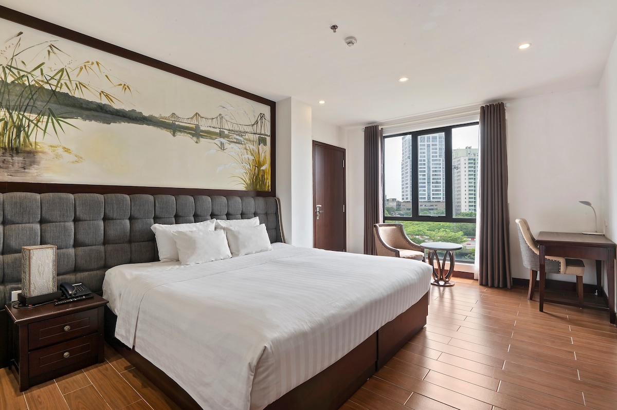 A modern bedroom features a spacious bed with a tufted gray headboard. Natural light filters through large windows, revealing a view of greenery and city buildings. A wooden desk and chair are positioned adjacent to the bed, complementing the soft color palette and decorative wall art.