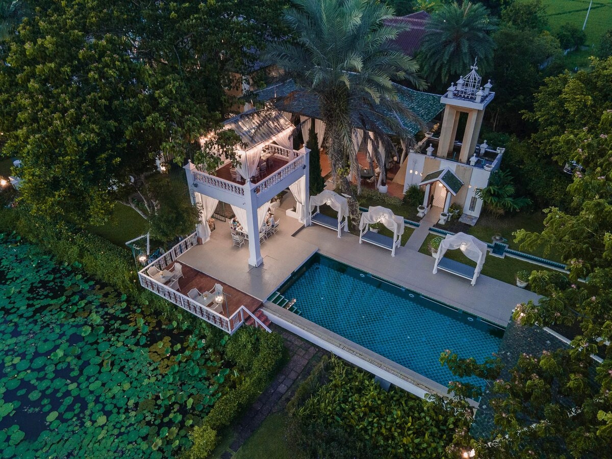 An aerial view of a villa surrounded by lush greenery, showcasing a large swimming pool and several relaxation pavilions. A tranquil lotus pond sits nearby, enhancing the serene outdoor atmosphere. The area is well-lit, indicating a welcoming space for leisure and relaxation.