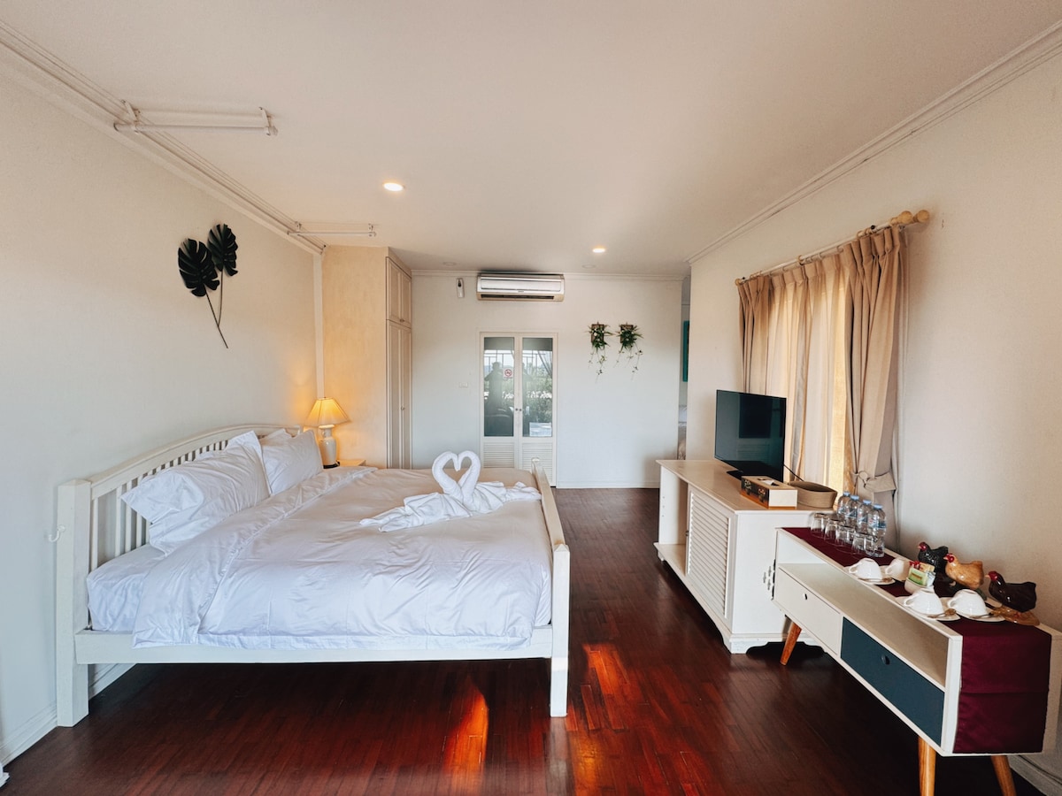The spacious room is furnished with a large bed, complemented by a decorative arrangement of towels shaped into a heart. Natural light filters through two windows, while a television and a side table are positioned against the wall. Wood flooring contributes to a warm ambiance.