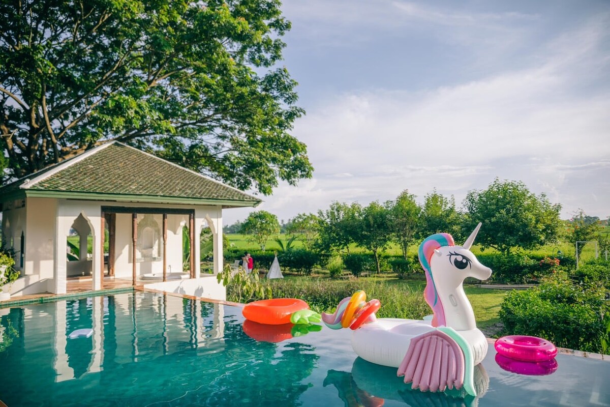 A tranquil swimming pool reflects the serene surroundings, featuring a colorful unicorn float and additional inflatable rings. Lush greenery frames the area, with a charming building visible in the background, creating an inviting atmosphere for relaxation.