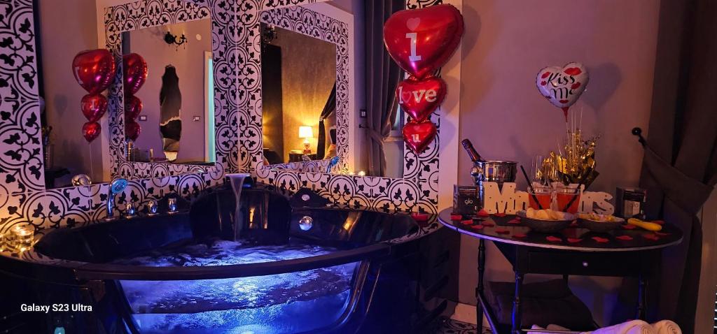 A dark, elegant bathroom features a large jacuzzi tub illuminated with soft blue lighting. Two decorative mirrors with intricate patterns enhance the space. The area is adorned with romantic details, including heart-shaped balloons and a tray with drinks, creating a festive ambiance.