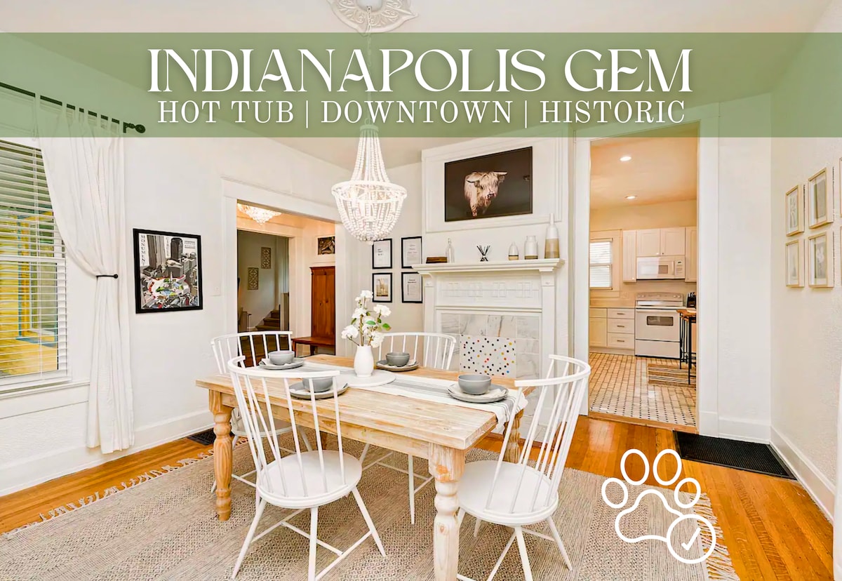 A charming dining area is presented with a wooden table set for six, complemented by white wire chairs. Natural light floods the space through large windows, and a decorative chandelier hangs above, providing an inviting atmosphere. The adjacent living area is visible, showcasing a cozy fireplace.