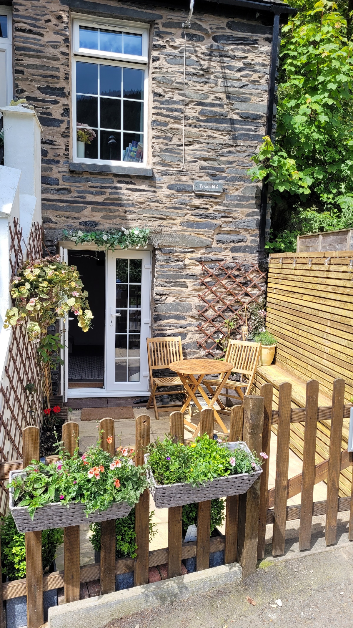 A private patio area is framed by a wooden fence and adorned with flower boxes. Wooden outdoor furniture, including a round table and chairs, invites relaxation. French doors lead into the cottage, complemented by climbing plants and natural stone wall textures.