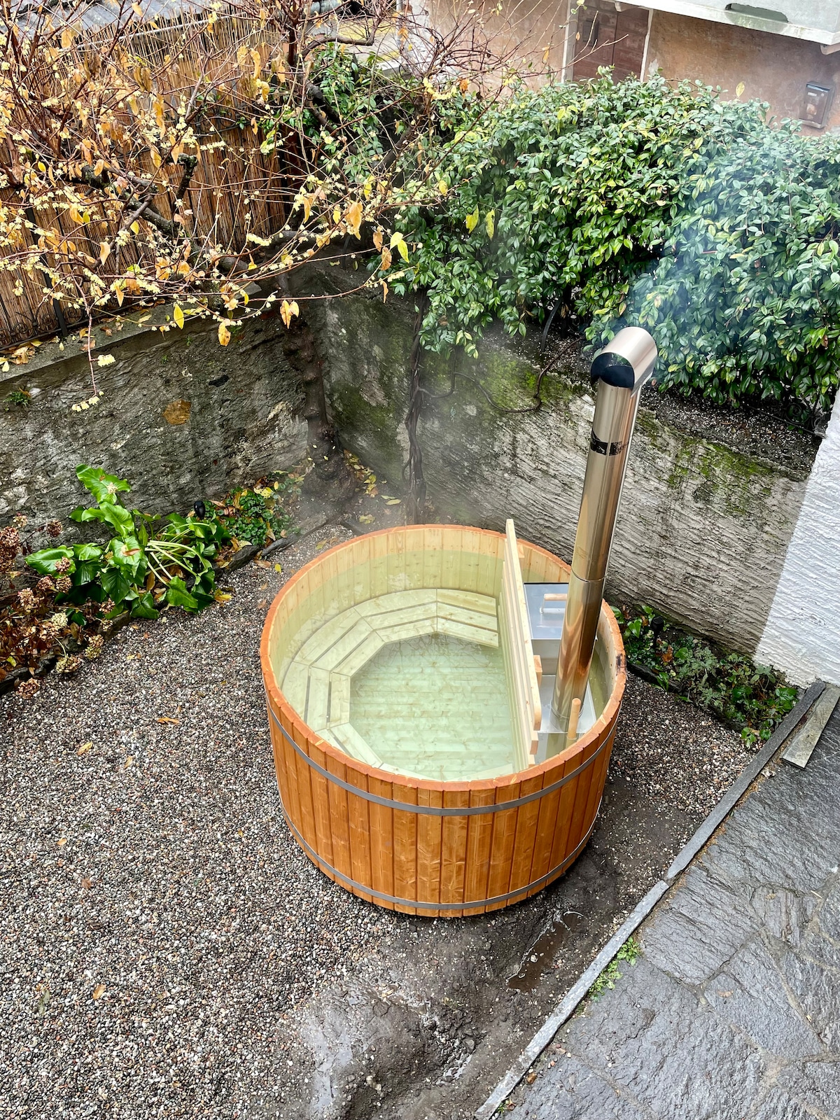 A wooden hot tub is situated in a gravel area, surrounded by plants and greenery. Steam rises gently from the heated water, while the metal chimney extends upward. The natural setting offers a serene backdrop for relaxation.