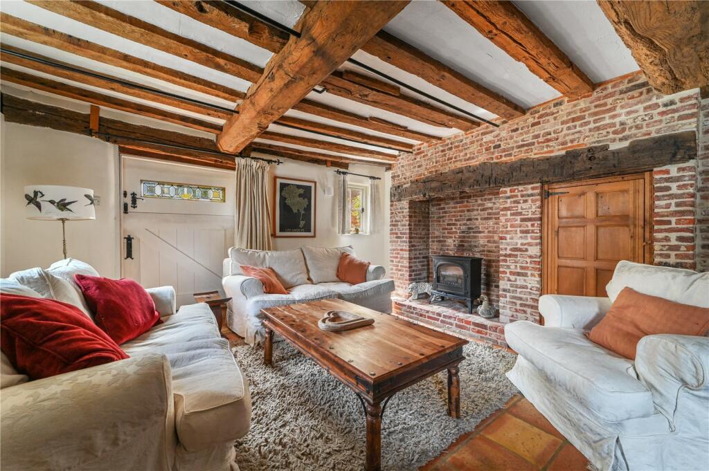 A cozy lounge features exposed wooden beams and a brick fireplace as its focal point. Two white sofas provide comfortable seating, complemented by a wooden coffee table at the center. Natural light enters from a window with stained glass, enhancing the warm ambiance.