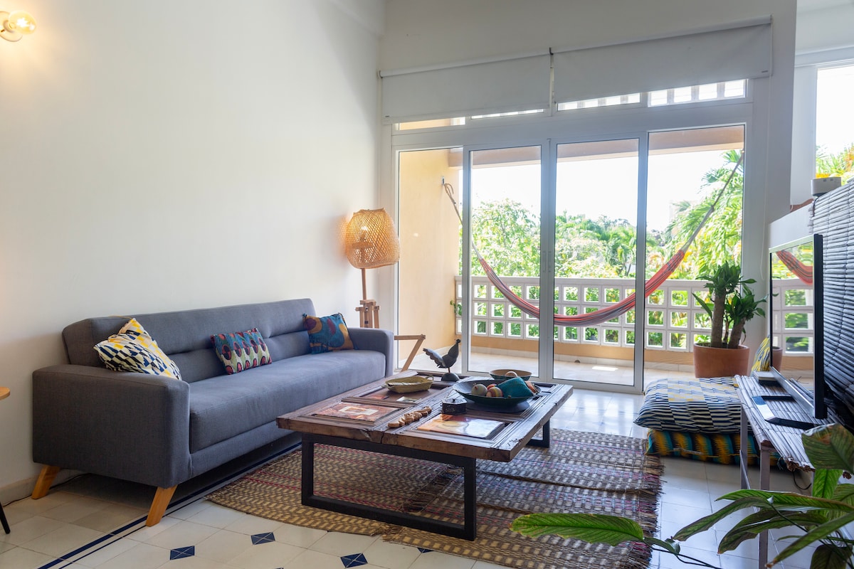 An open-plan living area features a comfortable grey sofa adorned with patterned cushions. A wooden coffee table sits centrally, flanked by a decorative rug. Floor-to-ceiling glass doors lead to a balcony, showcasing a hammock and views of the lush vegetation outside.