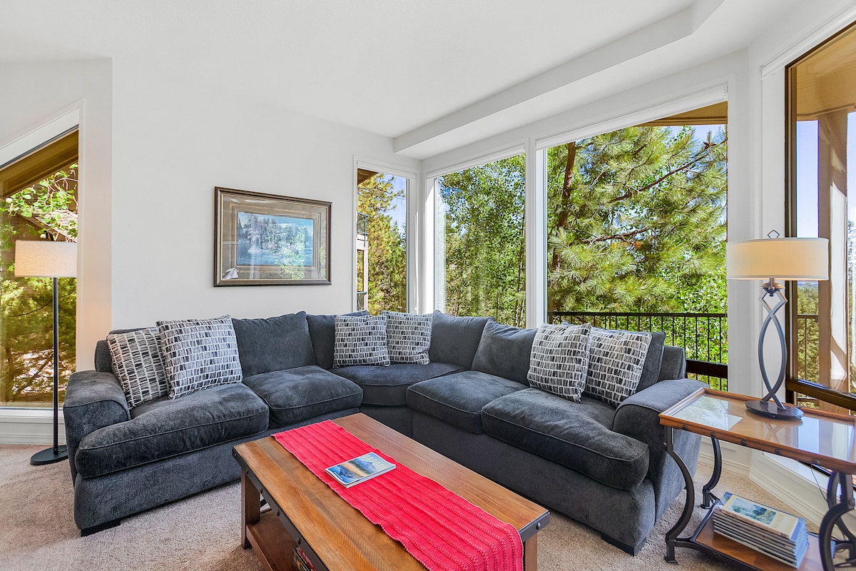 A spacious living room is highlighted by a large sectional sofa adorned with patterned pillows. Natural light fills the room through expansive windows, offering views of the surrounding trees. A wooden coffee table is positioned at the center, complemented by a side table with a lamp.