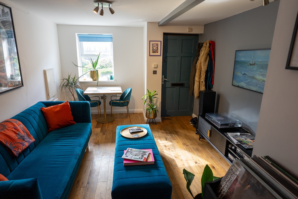 A comfortable living room features a vibrant blue sofa with colorful cushions. A small dining table is positioned near a window, accompanied by two teal chairs. A record player sits beside a collection of vinyl records, and a plant adds a touch of greenery.