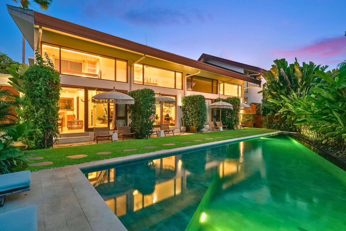 A spacious outdoor area is shown with a shimmering swimming pool surrounded by lush greenery. Comfortable lounge chairs and shaded umbrellas are placed on the deck. The modern structure behind features large glass windows, offering views into the inviting interior spaces.