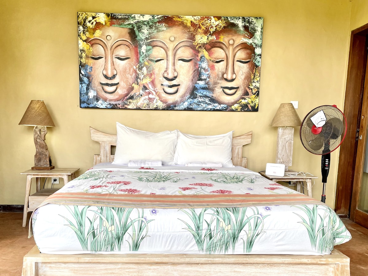 A spacious bed is positioned in the center of the room, adorned with a floral-patterned cover. Two bedside lamps flank the bed, and a fan is placed nearby. A colorful wall art piece in a wooden frame adds a touch of creativity to the warm-colored walls.