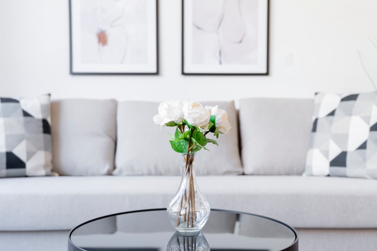 A modern living space features a light grey couch adorned with patterned cushions. A glass table sits in front, displaying a vase with white flowers, while abstract wall art is framed and hung above the couch.