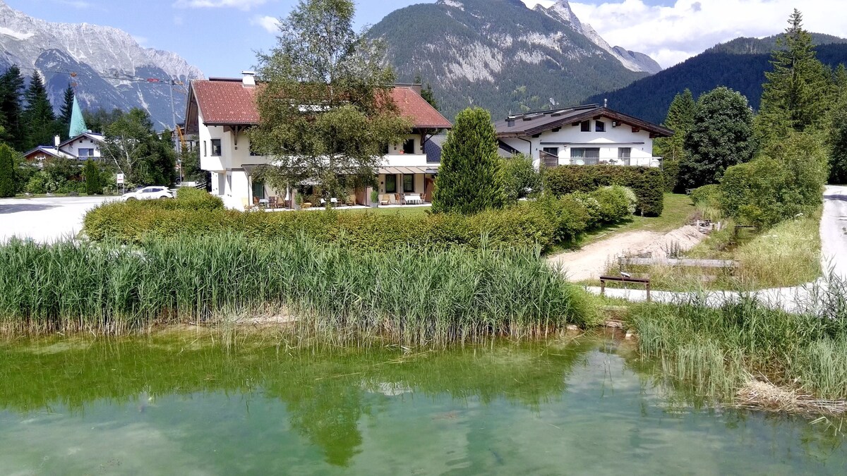 The image depicts a two-story building surrounded by greenery and water, with mountains rising in the background. A clear lake reflects the sky and is bordered by tall grass. The structure features large windows and a welcoming entrance, enhancing the natural setting.