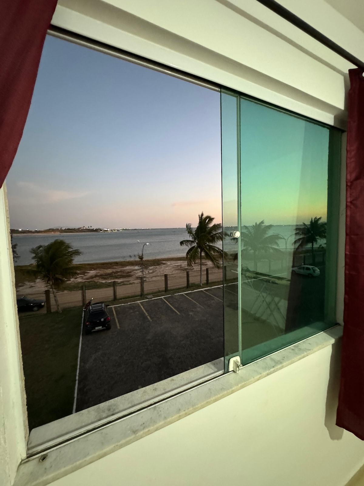 A view from a window showcases a serene lakeside scene, featuring palm trees swaying gently against the horizon. The tranquil water reflects soft hues of twilight, with parked cars visible along the water's edge.