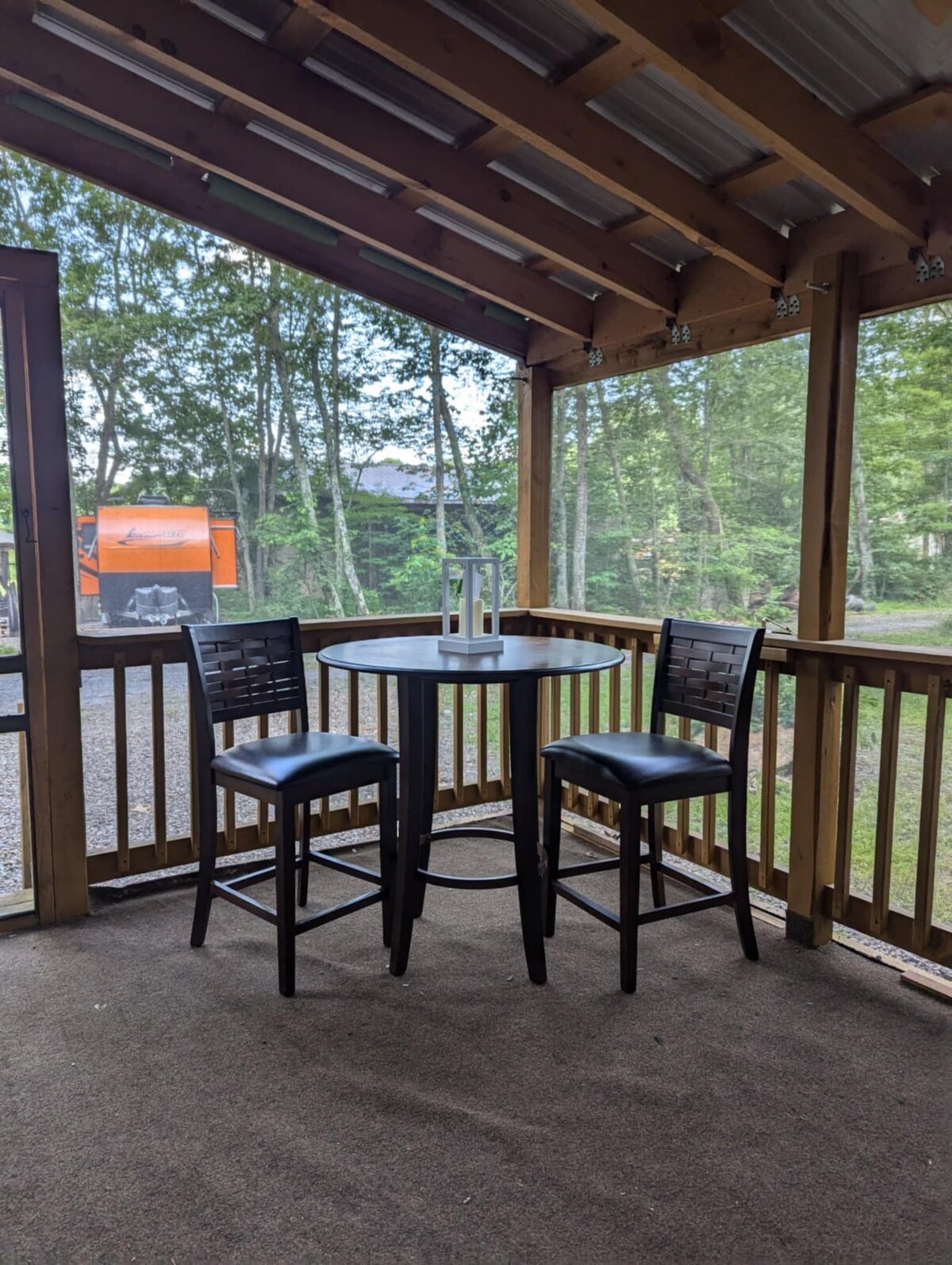 A small covered porch features a round table with two high chairs, providing a comfortable spot for conversation. Surrounded by wooden railings, the area is illuminated by natural light and overlooks a wooded landscape.