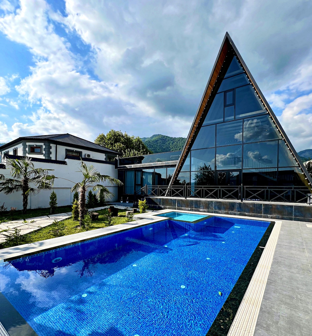 A contemporary chalet features a striking A-frame structure with large glass windows, reflecting the surrounding landscape. A spacious outdoor area includes a heated swimming pool, bordered by lush greenery and palm trees, creating a serene environment. The backdrop showcases picturesque mountains under a partly cloudy sky.