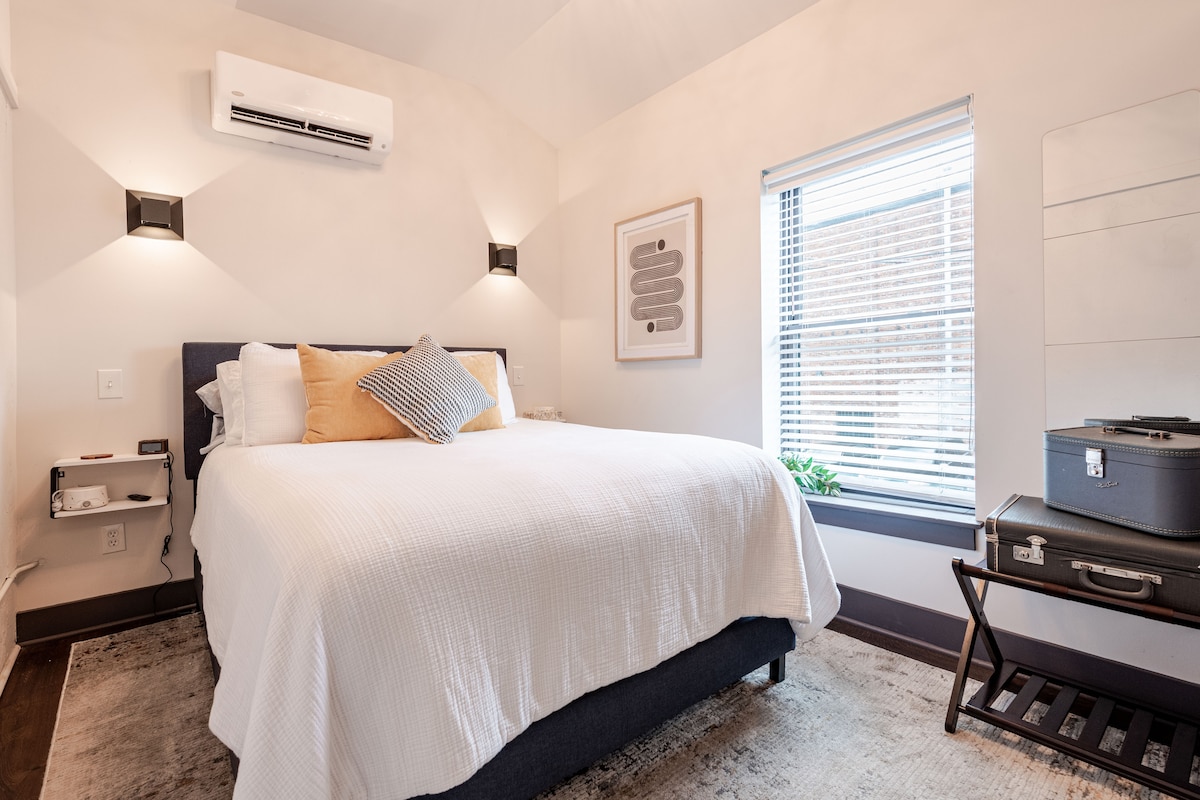 A cozy bedroom features a spacious bed dressed in crisp white linens, complemented by an array of decorative pillows. Natural light filters through a window adorned with blinds, illuminating the room's neutral tones. A small bedside table and a vintage suitcase add functional charm.