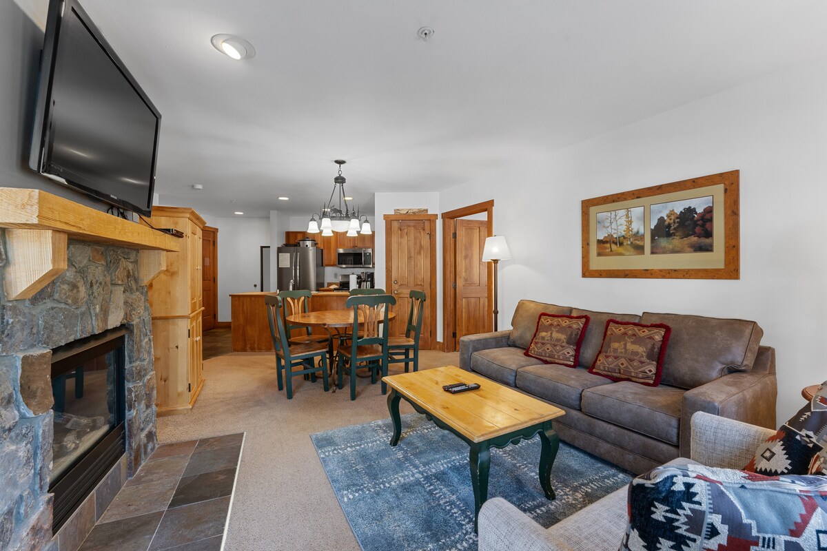 A living area features a comfortable sofa in neutral tones, complemented by decorative throw pillows. A flat screen TV is mounted above a stone fireplace. A dining table with seating for four is visible in the background, alongside a fully equipped kitchen.