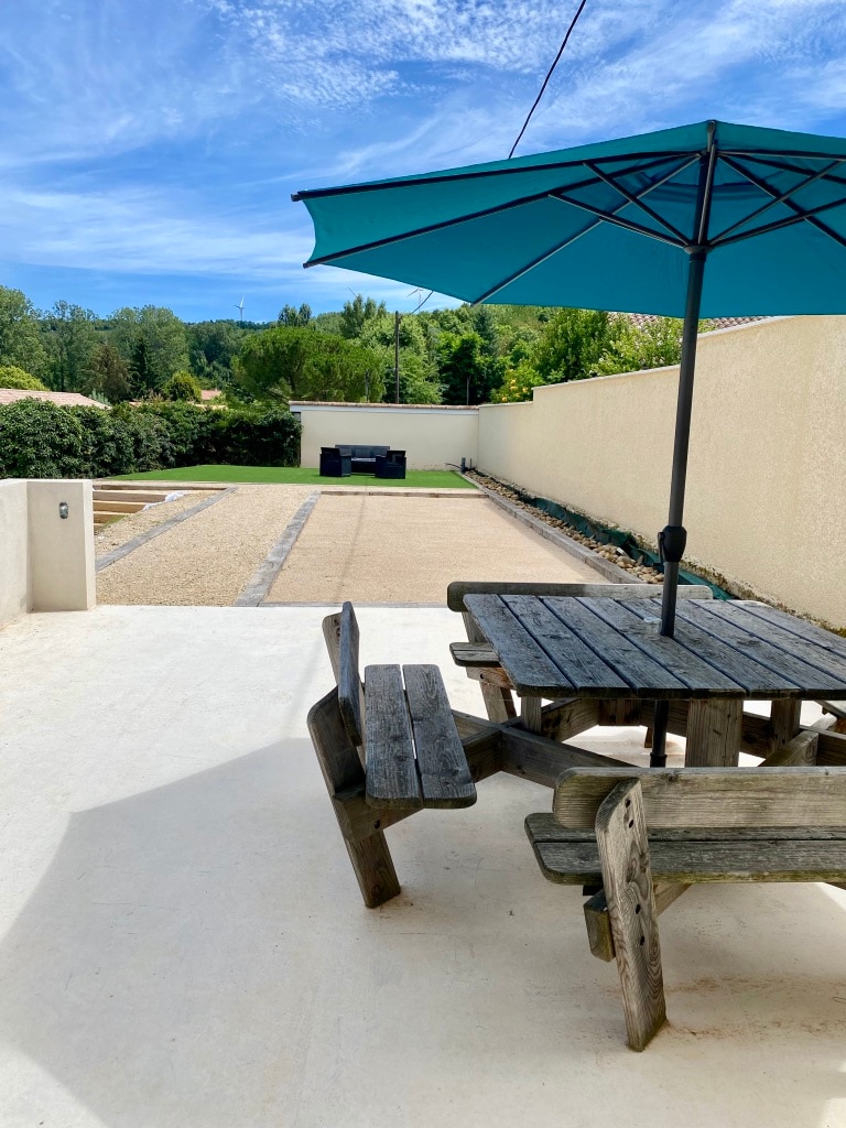 An outdoor seating area is furnished with a wooden table and benches under a large umbrella. A gravel area is visible in the background, with a petanque court located further back. Greenery and low hedges border the space.