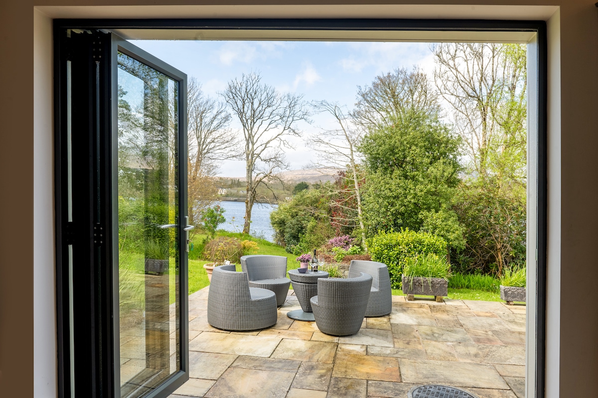 A patio area is visible through an open set of double doors, featuring modern, gray outdoor seating arranged around a small table. Lush greenery and trees frame the view of a tranquil body of water in the distance, contributing to a serene outdoor setting.