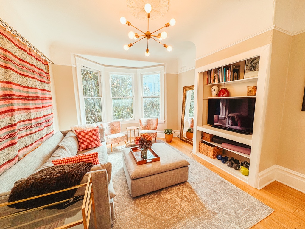 A light-filled living area features a large sectional sofa with patterned pillows, a coffee table in the center, and a cozy rug beneath. Decorative shelving holds books and art, while large windows provide views of greenery outside. A floor mirror adds a sense of space.
