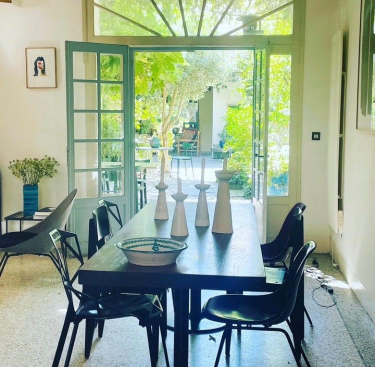 A dining area features a large wooden table set with decorative vases. The space is brightened by large glass doors that open to a lush garden, creating a seamless connection between indoor and outdoor settings.