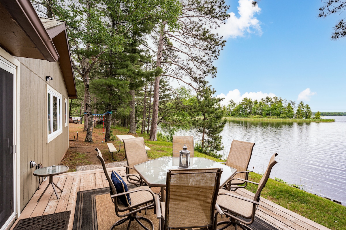An outdoor dining area is set on a wooden deck overlooking the lake. The space features a round table surrounded by six chairs, with a lantern centerpiece. Lush greenery and water create a peaceful backdrop, enhancing the inviting atmosphere.