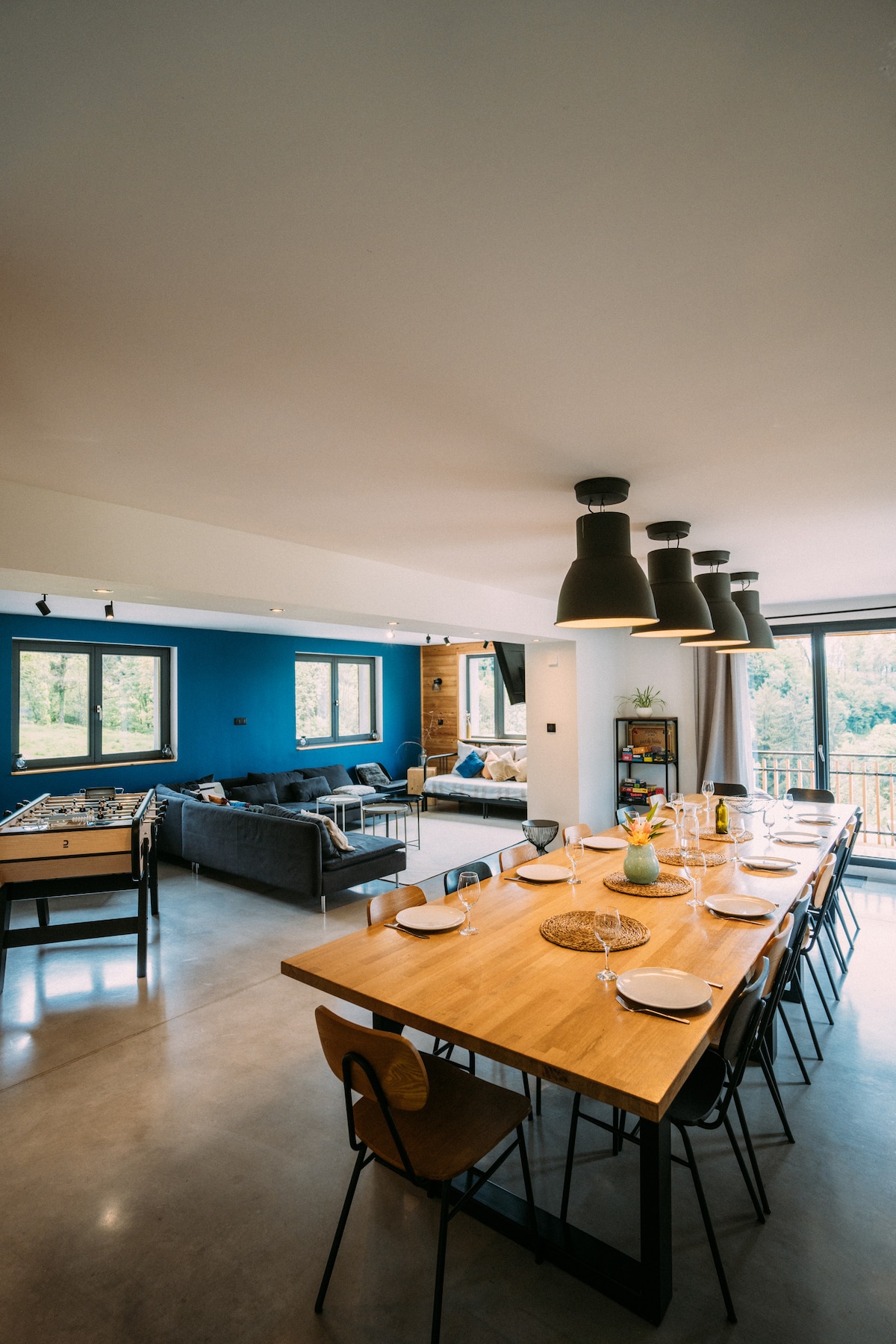 The open dining area features a large wooden table, set for multiple guests, with modern black chairs arranged around it. A comfortable seating space with a gray sofa and vibrant wall color is visible in the background, complemented by large windows that provide natural light.