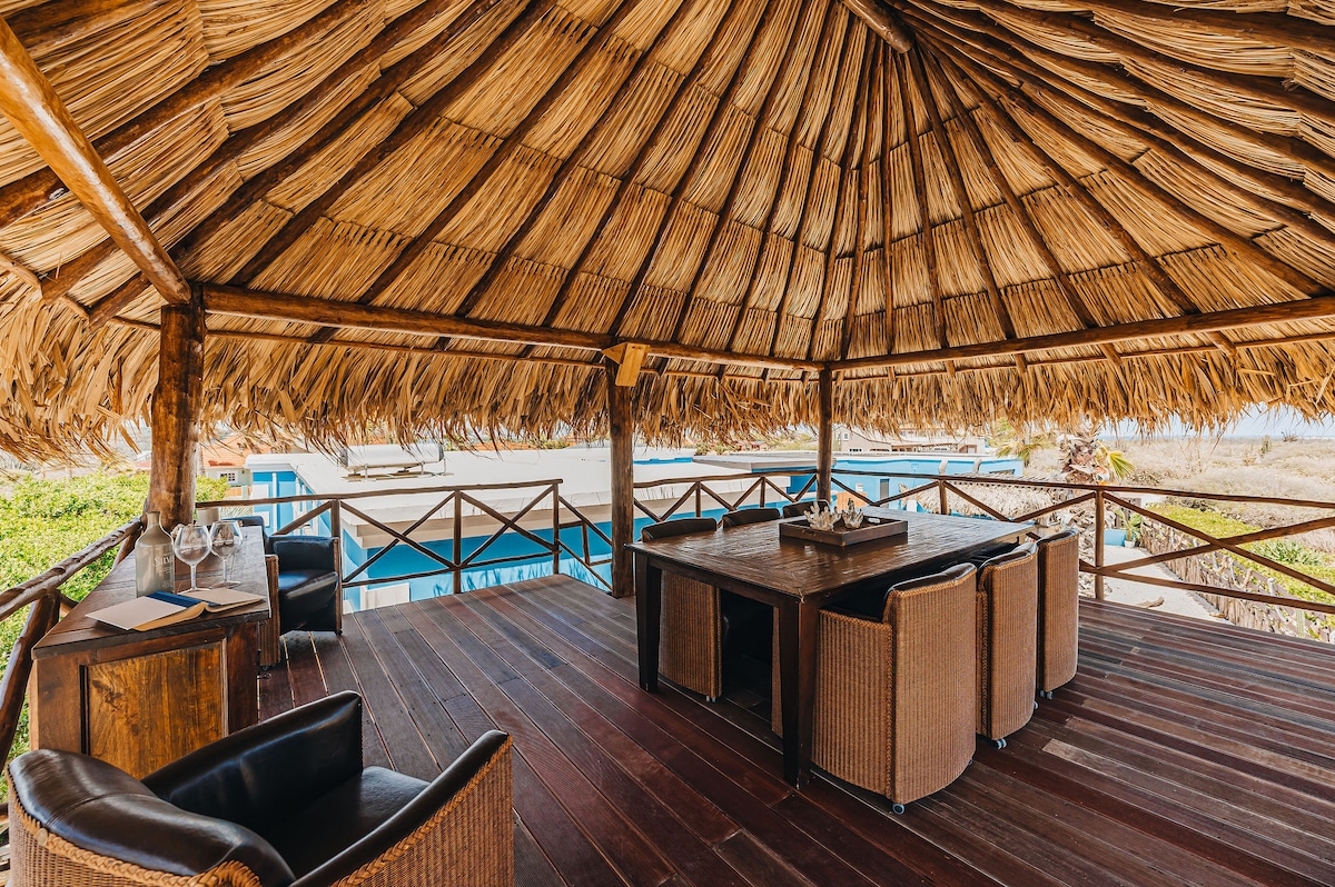An outdoor palapa features a thatched roof, providing shade over a spacious dining area with a large wooden table surrounded by woven chairs. The open design offers views of the surrounding landscape, creating a serene space for gatherings and meals.