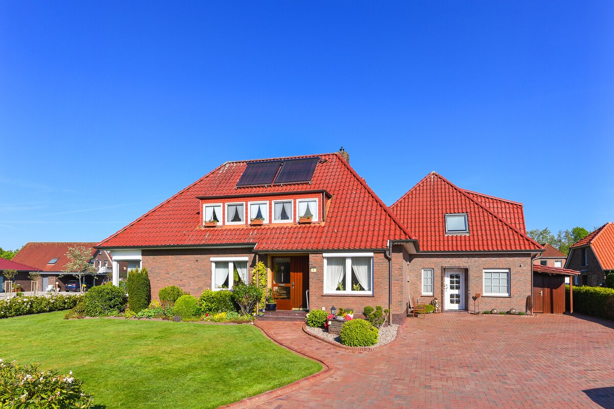 A spacious two-story house is set amidst a well-maintained garden. The exterior features a red-tiled roof and large windows, allowing natural light inside. A brick pathway leads to the entrance, while green shrubs and flower beds enhance the inviting landscape.