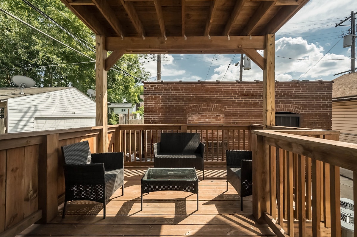 A wooden patio features a covered sitting area, furnished with four black wicker chairs surrounding a glass-topped table. The space offers a view of nearby trees and structures, creating a relaxing outdoor retreat.