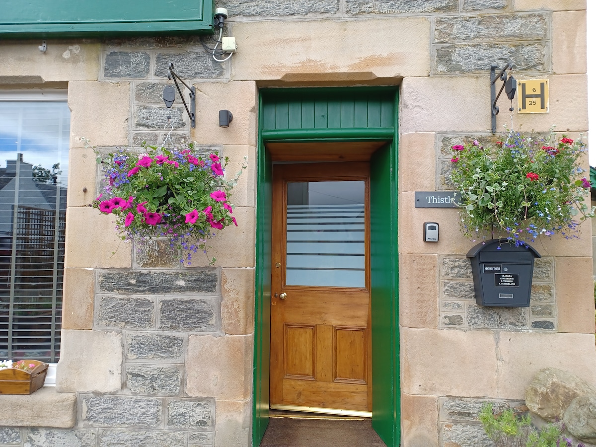 The entrance features a wooden door framed by flower baskets filled with vibrant pink and purple blooms. The stone exterior is complemented by clear glass panels in the door, allowing natural light to enter, while a mailbox is positioned to the right.