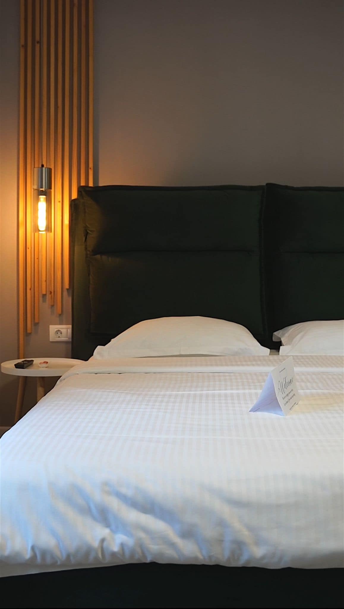 A cozy bedroom features a spacious bed with a simple white striped blanket. A green upholstered headboard provides a modern touch, while a small bedside table holds a welcome note. Soft lighting from a wall fixture creates a calm ambiance.