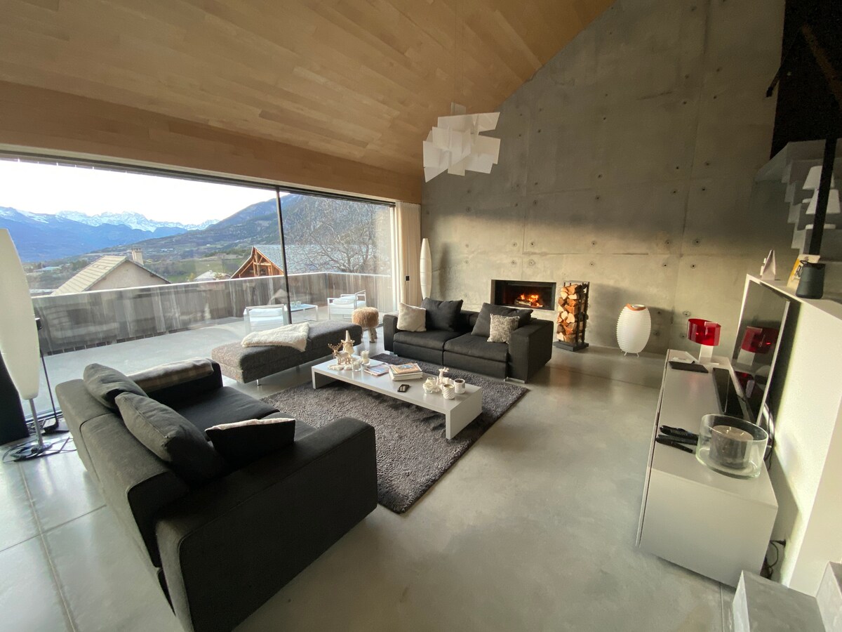 A spacious living area features large windows that showcase mountain views. Dark sofas are arranged thoughtfully on a soft rug, with a minimalist coffee table positioned at the center. A modern fireplace adds warmth to the room, enhanced by warm tones from unique light fixtures.