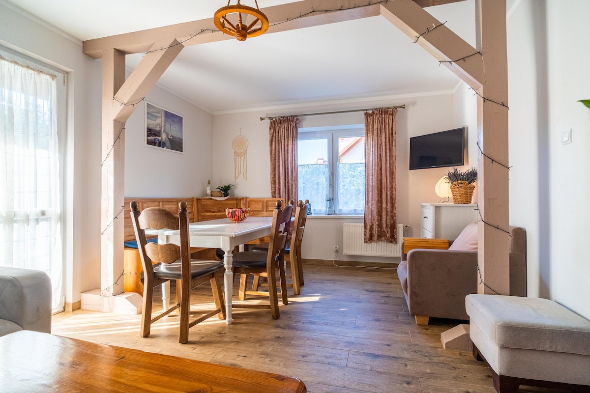 A spacious dining area is framed by a wooden arch, featuring a large table surrounded by six chairs. Large windows provide natural light, complementing the light-colored walls and wood flooring. A cozy seating area is visible in the corner, alongside a flat-screen television.