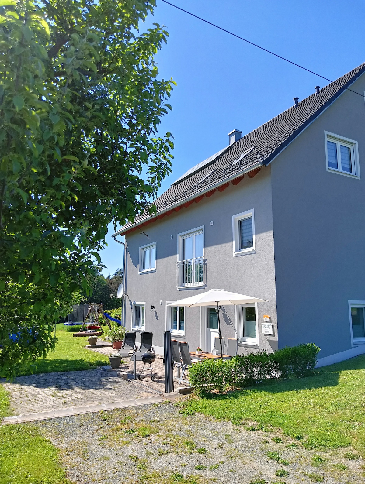 The exterior of a two-story gray house is shown, featuring a well-maintained garden with a lawn. A patio area is furnished with chairs and an umbrella, situated near a tree. The layout suggests a welcoming space for relaxation and outdoor activities.