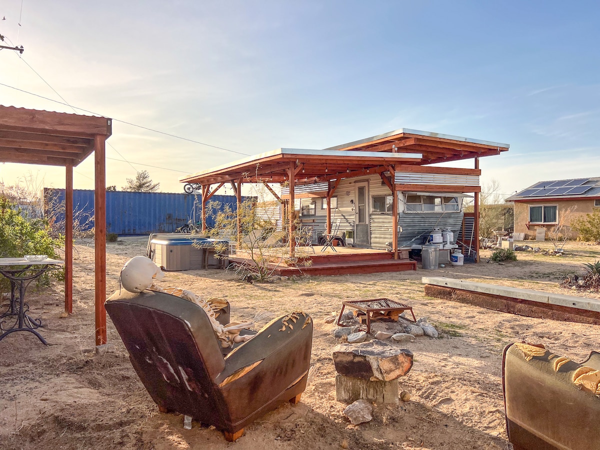 The exterior of the RV is framed by a spacious wooden deck and features a hot tub surrounded by desert landscaping. Cozy seating arrangements, including armchairs and a fire pit, create an inviting outdoor atmosphere. The sky is clear, emphasizing the tranquil desert setting.