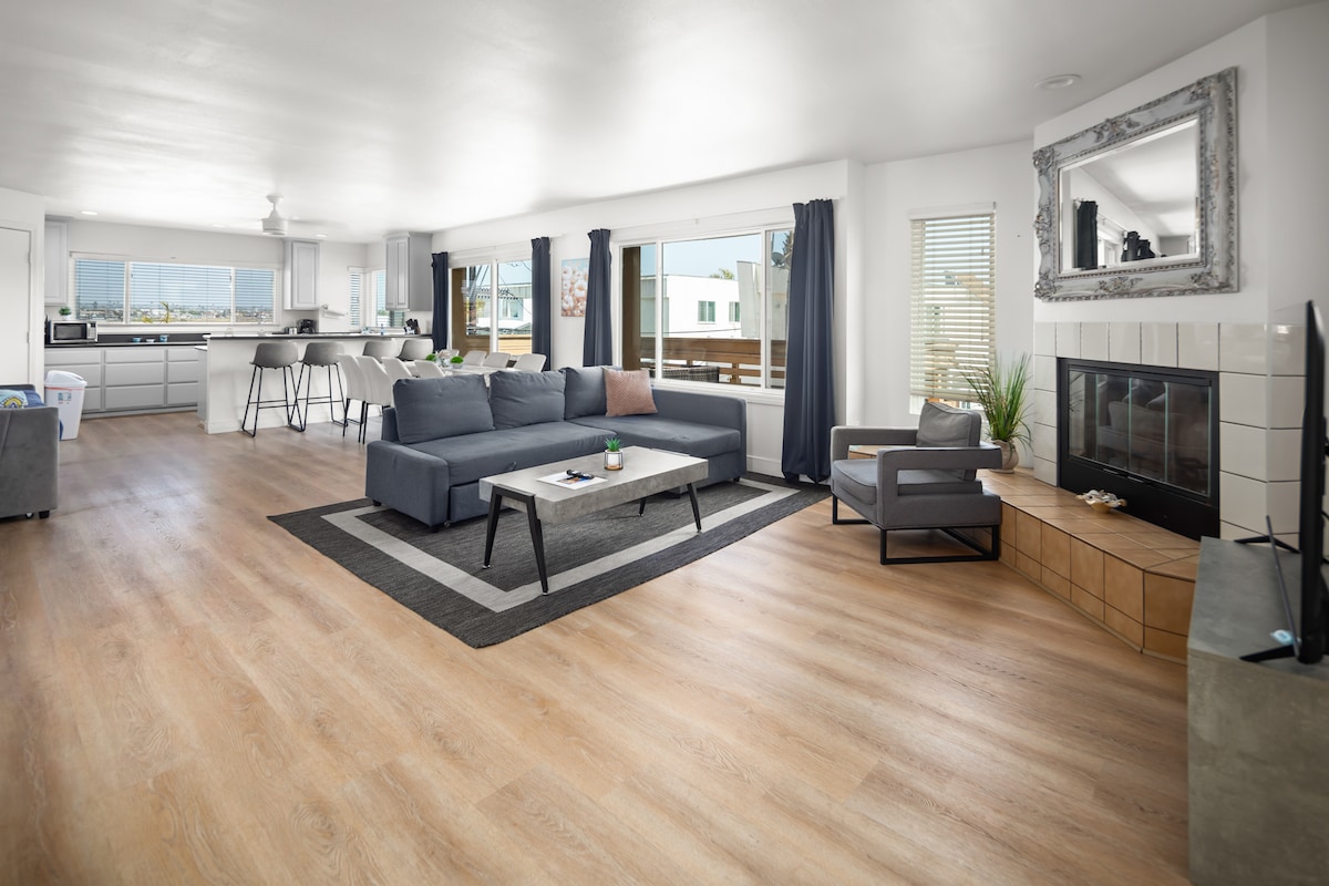An inviting living area features a spacious sectional sofa paired with a stylish armchair. Natural light streams through large windows, illuminating the wood-style flooring. A modern fireplace and decorative mirror add subtle elegance, while the open floor plan flows into the adjacent kitchen.