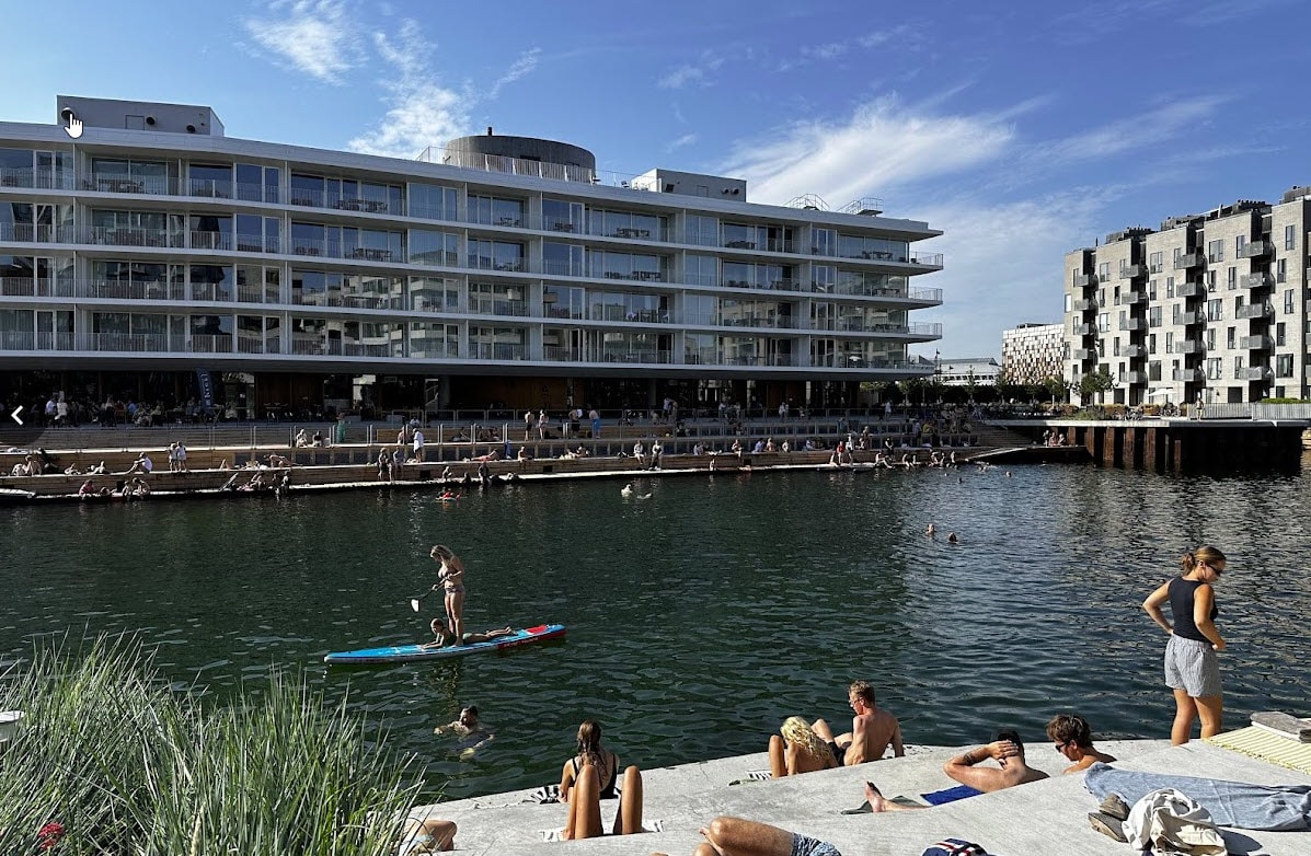 A waterfront scene captures a modern multi-story building with extensive glass facades, reflecting sunlight. Individuals can be seen enjoying water activities and relaxing along the inviting shoreline. Lush greenery frames the concrete steps leading into the water, enhancing the outdoor setting.
