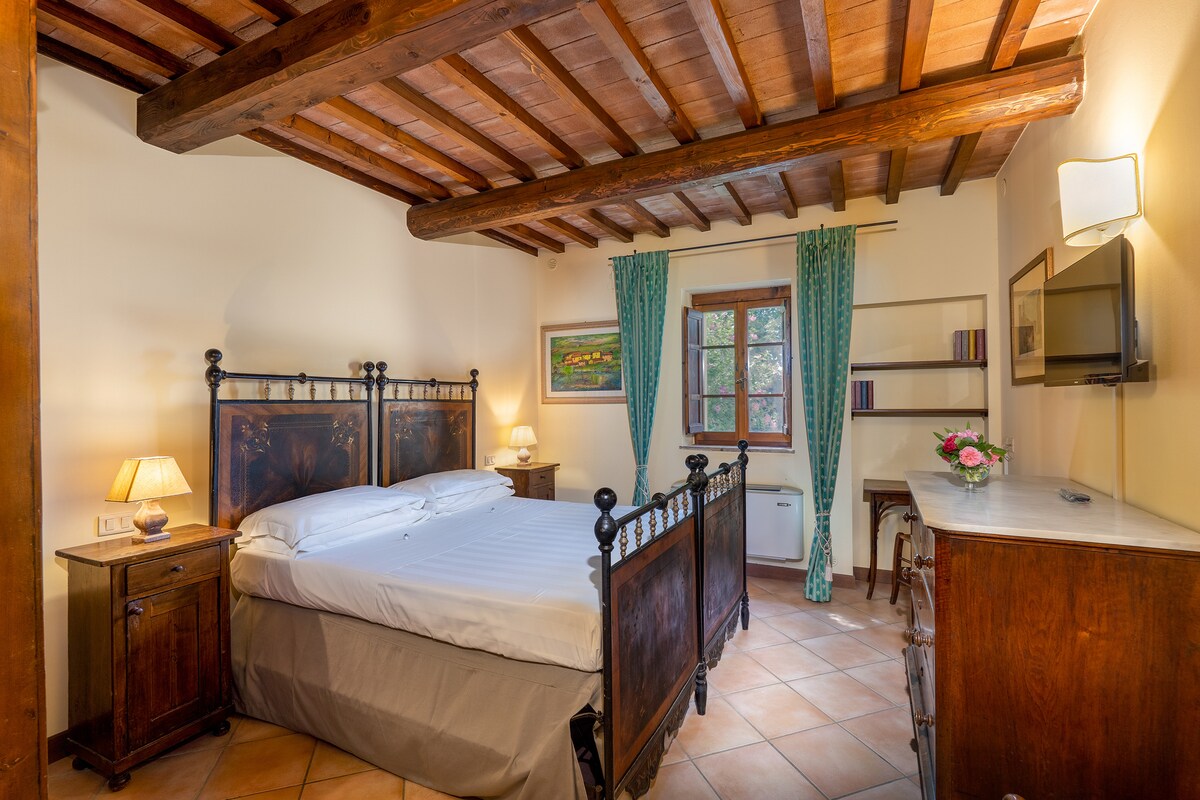 A spacious bedroom is adorned with rustic wooden beams on the ceiling. A large bed with a dark wooden frame is positioned in the center, accompanied by bedside lamps. A window with turquoise drapes offers natural light, while a wooden dresser and shelf hold decorative items.