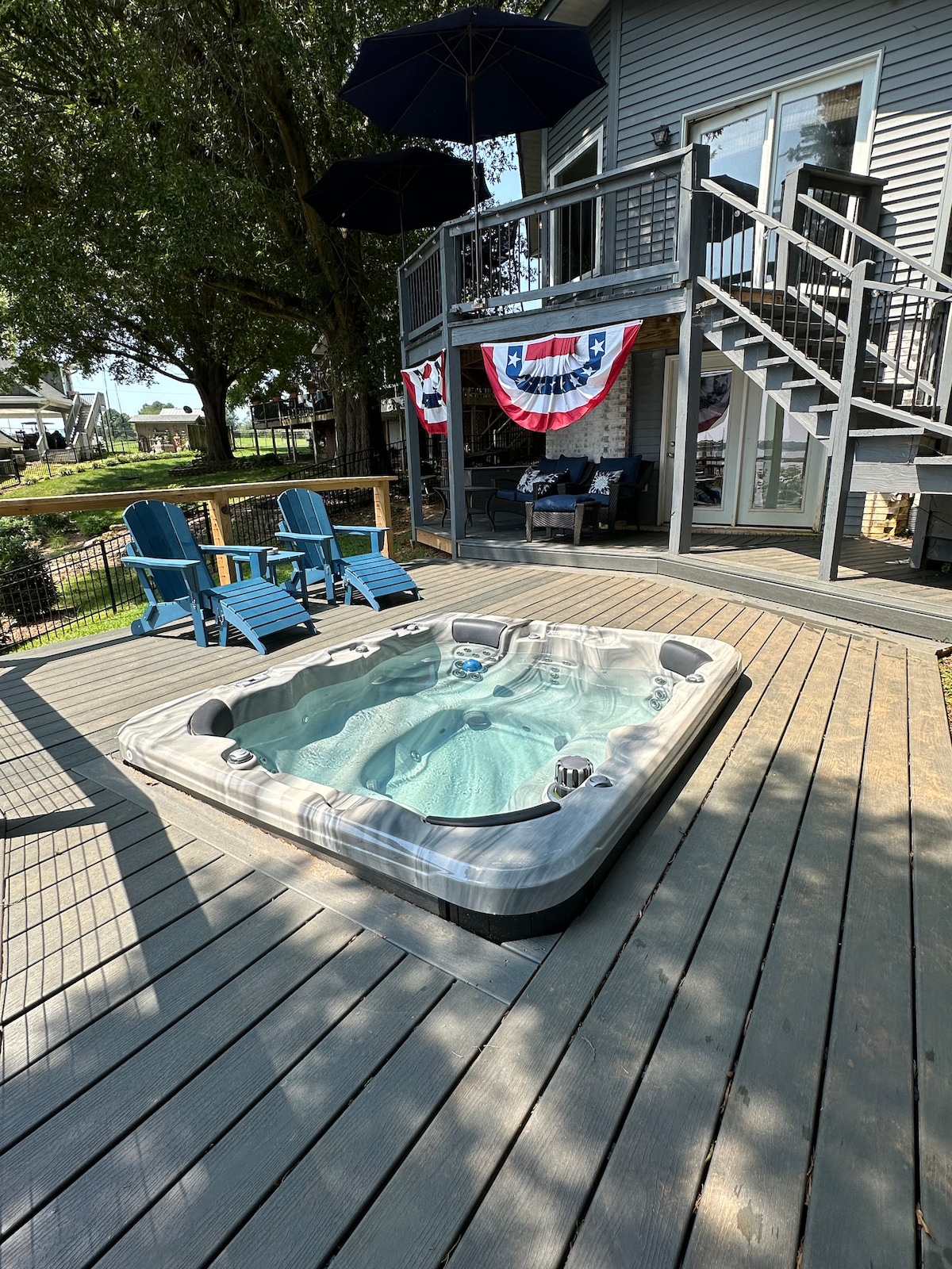 A hot tub accommodates seven people, positioned on a Trex deck. Two blue lounge chairs are situated nearby, and a staircase leads to the upper level of the home. A decorative red, white, and blue banner is displayed, adding a touch of color to the serene outdoor setting.