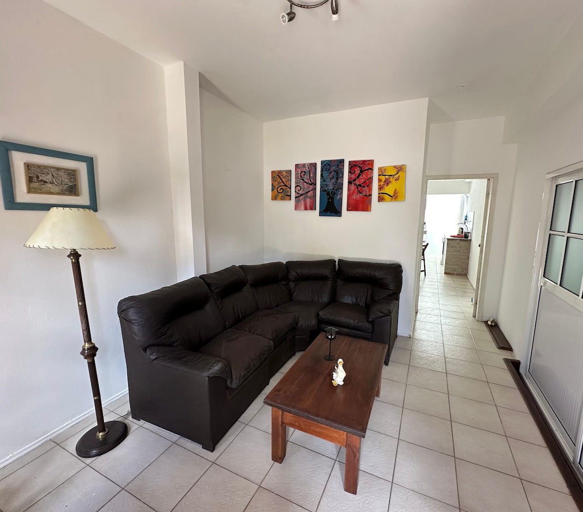 A comfortable living area is displayed, featuring a dark leather sectional sofa arranged around a wooden coffee table. Wall art in vibrant colors decorates the space, while a standing lamp provides additional lighting. A doorway leads to the kitchen area at the back.