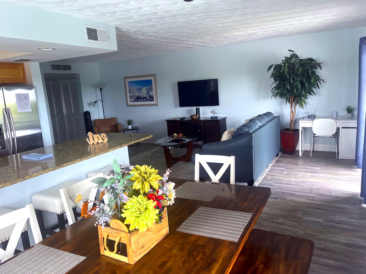 An open-concept living area features a spacious kitchen with a granite countertop and dining table. The cozy living room includes a plush sofa facing a flat-screen TV. A decorative plant and a flower arrangement add a touch of color, enhancing the inviting environment.