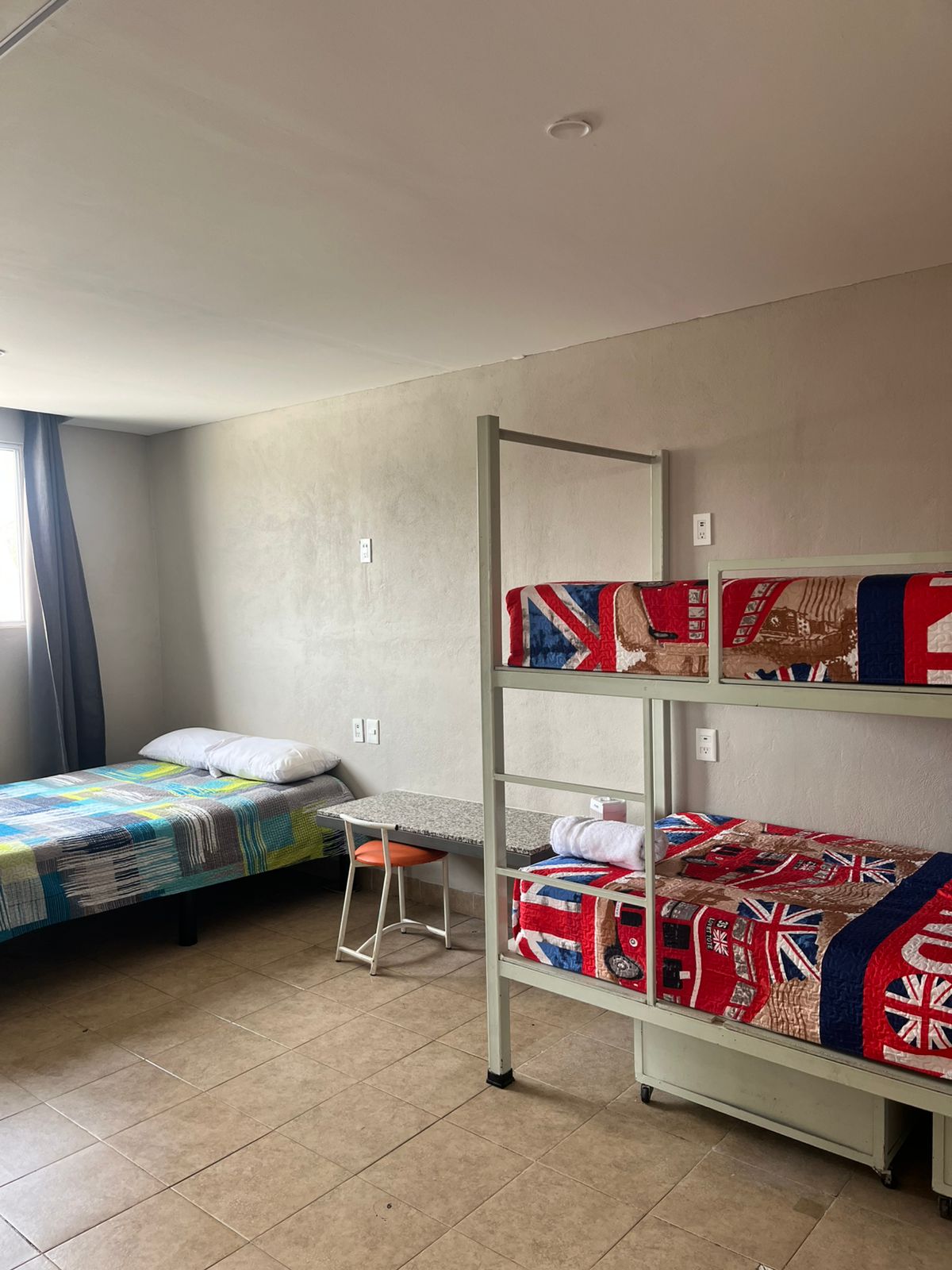 A simple guestroom features a spacious bed alongside a bunk bed with colorful bedding. A small table and stool are positioned nearby against the wall. Natural light filters in through a window, enhancing the light-toned walls and tiled floor.