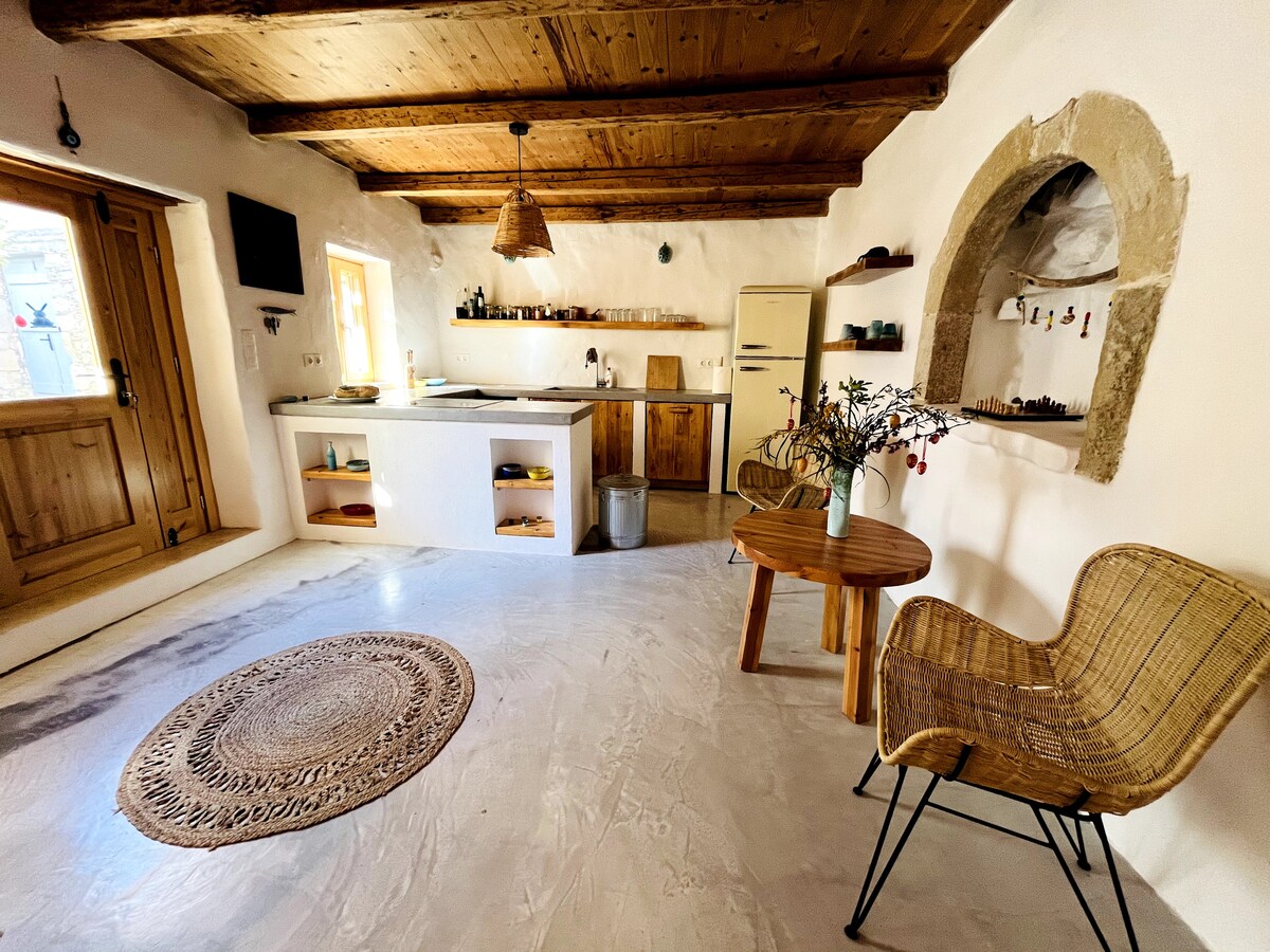 The spacious kitchen area features a large island with natural wood accents, complemented by wooden shelves and an antique-style refrigerator. A round table and woven chair create a cozy seating space, while a textured rug enhances the earthy tones of the room, illuminated by natural light from the window.